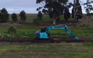 Blue excavator removing old rail lines in green field on overcast day at Coldstream