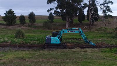 Blue excavator removing old rail lines in green field on overcast day at Coldstream